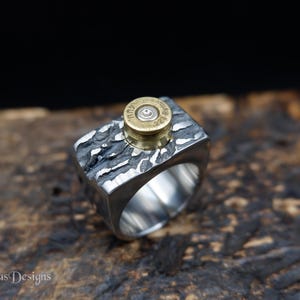 Peut inclure: Une bague argentée présentant une surface sombre, texturée et rugueuse, surmontée d'un culot de douille en laiton. Le culot porte l'inscription .223 REM. La bague possède un anneau large et poli avec une finition intérieure lisse.
