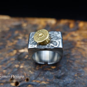 Op de afbeelding: Zilverkleurige ring met een rechthoekige bovenkant, met een getextureerd oppervlak en een goudkleurige kogelhuls. De ring heeft een brede band en een uniek, industrieel ontwerp. De ring is een statement-stuk.