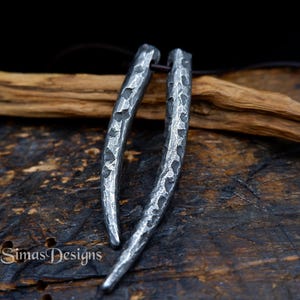 May include: Two silver metal pendants shaped like long, thin horns with a hammered texture. The pendants are hanging from a brown leather cord.