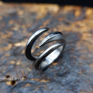 Op de afbeelding: Een donker zilverkleurige metalen ring met een open bypass-ontwerp en twee taps toelopende, puntige uiteinden. Het oppervlak van de ring heeft een getextureerde, gehamerde afwerking met zichtbare oxidatie in de groeven. De tekst SimasDesigns is zichtbaar.