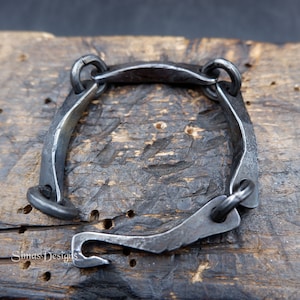 Op de afbeelding: Handgesmede armband van donker metaal, vierkant van vorm. De armband heeft een haak-oogsluiting en is verbonden door cirkelvormige schakels. Het metaal heeft een getextureerd, gehamerd uiterlijk, wat wijst op een rustiek, ambachtelijk ontwerp. De armband wordt gepresenteerd op een houten oppervlak.