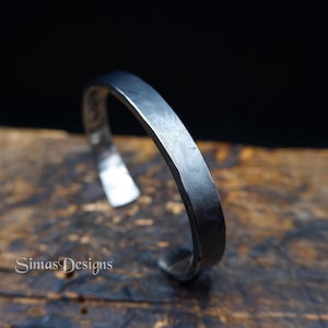 May include: A silver-coloured metal cuff bracelet with a hammered texture. The open-ended bracelet has a rectangular shape and is displayed on a wooden surface. The brand name "Simas Designs" is visible in the lower left corner.