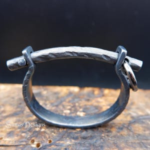 May include: A handcrafted, dark gray metal ring with a textured, hammered finish. The ring features a curved bar across the top, held in place by two square brackets. Two small silver rings hang from one side. The ring is a unique piece of jewelry.