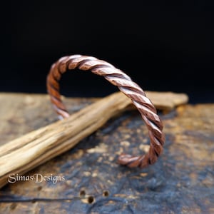May include: A copper-colored, twisted metal cuff bracelet. The bracelet has a textured, rope-like design. The jewelry is displayed on a wooden surface with a dark background. The bracelet is a handmade piece.