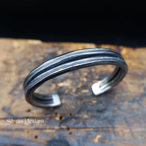 Op de afbeelding: Een zilverkleurige manchetarmband met een getextureerd, gelaagd ontwerp. De open manchet heeft een reeks parallelle lijnen, waardoor een gedurfde, industriële esthetiek ontstaat. De armband wordt gepresenteerd op een houten oppervlak.