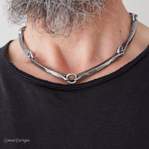 Puede incluir: Un collar de metal gris oscuro que presenta una serie de barras curvas con textura martillada conectadas por pequeños anillos circulares. El collar descansa sobre una camiseta negra, mostrando un diseño rígido y segmentado.