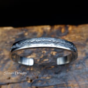 Könnte beinhalten: Ein silbernes Armreif-Armband mit einem strukturierten, bergähnlichen Design auf der Oberseite. Das offene Armband hat eine polierte, reflektierende Oberfläche. Der Markenname "Simas Designs" ist unten links sichtbar.