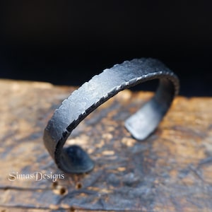 Op de afbeelding: Een donkergrijze metalen manchetarmband met een getextureerd, gehamerd oppervlak en ingekeepte randen. De armband heeft een gebogen, open ontwerp met een matte afwerking. De tekst SimasDesigns is zichtbaar in de linkerbenedenhoek.