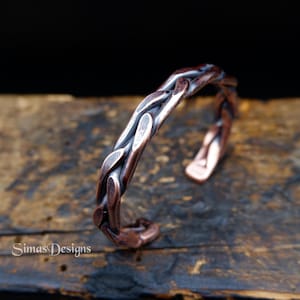 Op de afbeelding: Een koperen manchetarmband met een gevlochten ontwerp. De open manchet heeft een getextureerd, geweven patroon en wordt gepresenteerd op een houten oppervlak. De armband heeft een rustieke, handgemaakte uitstraling. De tekst "Simas Designs" is zichtbaar.