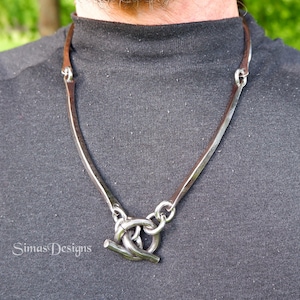 Op de afbeelding: Een zilverkleurige ketting met een bruin leren koord. De ketting heeft een uniek ontwerp met een tuimelsluiting. Het wordt gedragen op een donkergrijs shirt. De merknaam "Simas Designs" is links zichtbaar.