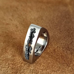 May include: A silver-toned metal ring with a smooth, polished finish and a wide, flat band. A vertical, jagged, dark grey textured inlay runs down the centre of the outer surface, creating a stark contrast against the metallic band.