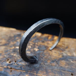 Op de afbeelding: Een handgemaakte, zilverkleurige manchetarmband met een getextureerde, gehamerde afwerking. Het open ontwerp heeft een vierkant profiel en wordt gepresenteerd op een houten oppervlak. Een minimalistisch accessoire.