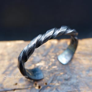 May include: A handcrafted, twisted metal cuff bracelet. The bracelet is made of dark, textured metal with a spiral design. The ends of the bracelet are slightly flared. The text "Simas Designs" is visible on the surface.