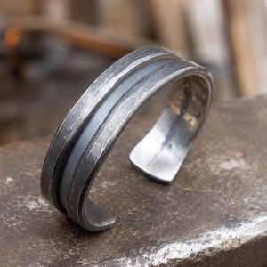 Puede incluir: Una pulsera brazalete de metal gris oscuro con un acabado martillado y texturizado en los bordes exteriores y un canal central liso y hundido. El diseño abierto muestra una apariencia oxidada y desgastada con una superficie mate.