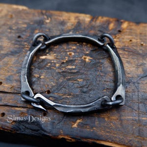 Op de afbeelding: Handgesmede, vierkante armband van donker, getextureerd metaal. De armband heeft een haak-en-oogsluiting en is verbonden door kleine, cirkelvormige schakels. De armband wordt getoond op een donker, houten oppervlak. De tekst "Simas Designs" is zichtbaar.