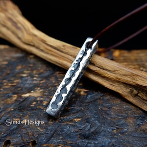 Op de afbeelding: Een zilverkleurige, rechthoekige hanger met een getextureerd oppervlak, hangend aan een bruin leren koord. De hanger heeft een robuuste, handgemaakte uitstraling. De achtergrond heeft een houten element, wat de rustieke esthetiek versterkt. De hanger is van Simas Designs.