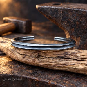 Könnte beinhalten: Ein dunkelgraues Armband aus Metall mit einer texturierten, gehämmerten Oberfläche und einem mittigen horizontalen Grat. Das Armband hat ein offenes Design und ein mattes Aussehen, das eine raue, industrielle Ästhetik mit sichtbarer Handwerkskunst zeigt.