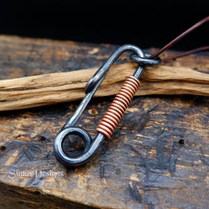 Può includere: Una collana con pendente artigianale in stile industriale. Il pendente ha la forma di una grande spilla da balia grigio scuro, con un dettaglio a spirale in filo di rame. È appeso a un cordino di cuoio marrone. Il marchio "Simas Designs" è visibile.
