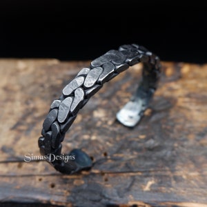 May include: A handcrafted, open-ended cuff bracelet with a textured, chain-like design. The bracelet is made of dark, oxidized metal, and the surface has a hammered appearance. The bracelet is displayed on a wooden surface.