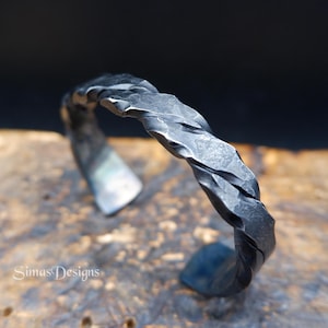 Puede incluir: Una pulsera de brazalete de metal gris oscuro con un diseño trenzado y texturizado con un acabado martillado. La superficie del metal muestra facetas irregulares y una apariencia mate. El texto SimasDesigns es visible en la esquina inferior izquierda.