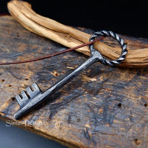 Puede incluir: Colgante de llave de metal oscuro hecho a mano con un detalle de anillo retorcido. La llave tiene un acabado texturizado y un cordón de cuero. El colgante descansa sobre una superficie de madera rústica con un trozo de madera flotante.