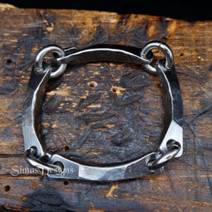 Op de afbeelding: Een handgemaakte, vierkante armband van gehamerd metaal. De armband heeft vier afgeronde verbindingsringen en een getextureerde, donkergrijze afwerking. Het ontwerp is industrieel en rustiek, geschikt voor dagelijks gebruik.
