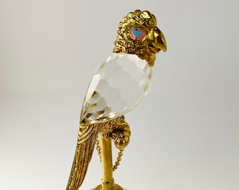 May include: A crystal parrot figurine with a gold-colored perch and base. The parrot's head is detailed with gold and features a blue and pink eye. The body is made of clear, faceted crystal.