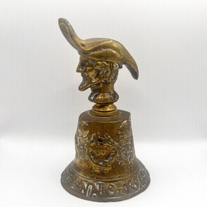 May include: Antique brass bell with a detailed design. The bell features a jester's head at the top and ornate relief patterns around the bell's body. The base of the bell has the word "WAGNER" engraved.