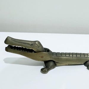 May include: A brass alligator nutcracker with an open mouth, detailed teeth, and a textured body. The alligator is resting on four small feet. The nutcracker is a decorative kitchen tool.