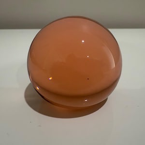 May include: A spherical, orange-tinted glass object. The smooth, glossy surface reflects light, creating highlights. The object sits on a white surface, casting a shadow. The overall shape is a perfect sphere.