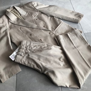 Puede incluir: Un traje beige de doble botonadura, que incluye una chaqueta y un pantal&oacute;n. La chaqueta tiene seis botones y solapas con muescas. El pantal&oacute;n tiene pliegues y pernera recta. El traje es de tejido liso y est&aacute; sobre una superficie embaldosada.