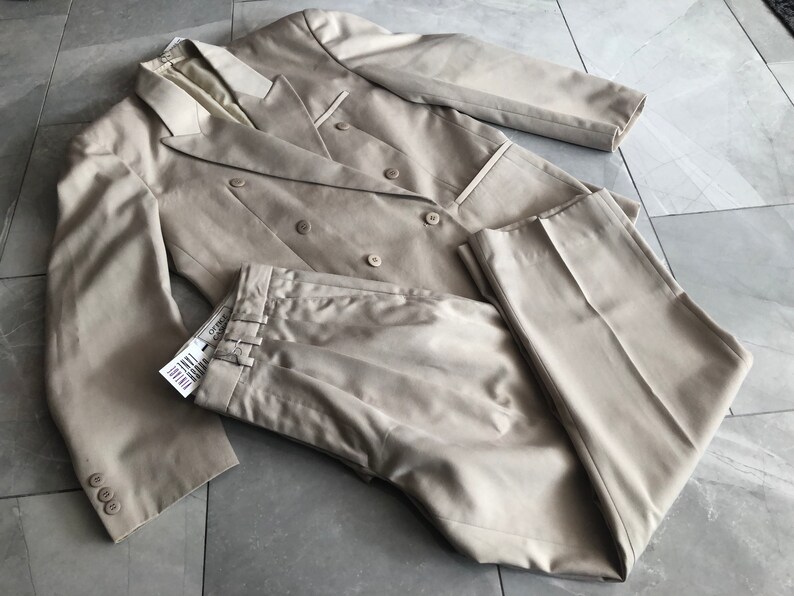 Puede incluir: Un traje beige de doble botonadura, que incluye una chaqueta y un pantal&oacute;n. La chaqueta tiene seis botones y solapas con muescas. El pantal&oacute;n tiene pliegues y pernera recta. El traje es de tejido liso y est&aacute; sobre una superficie embaldosada.