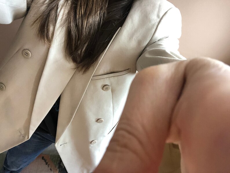 Puede incluir: Un blazer beige de doble botonadura con cuatro botones y solapas con muescas. El blazer tiene un bolsillo en el lado izquierdo y se usa sobre una camisa oscura y vaqueros azules. El blazer es de color neutro y parece estar hecho de una tela ligera.