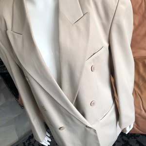 Puede incluir: Un blazer beige de doble botonadura con solapa de muesca cl&aacute;sica y dos filas de botones. El blazer tiene un corte entallado y un bolsillo en el lado izquierdo. La prenda se exhibe en un maniqu&iacute; blanco.
