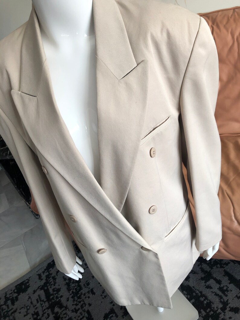 Puede incluir: Un blazer beige de doble botonadura con solapa de muesca cl&aacute;sica y dos filas de botones. El blazer tiene un corte entallado y un bolsillo en el lado izquierdo. La prenda se exhibe en un maniqu&iacute; blanco.