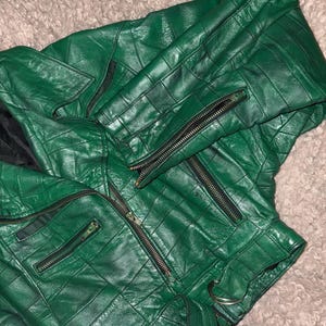 Peut inclure: Veste en cuir vert vif avec un motif matelassé. La veste comporte plusieurs poches zippées et une ceinture. Les fermetures éclair sont noires et le cuir a un léger éclat. La veste est pliée, mettant en valeur son design.