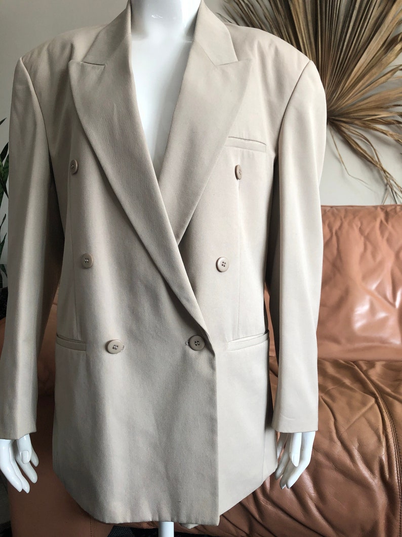 Puede incluir: Un blazer beige de doble botonadura con cuatro botones a juego y dos bolsillos delanteros. El blazer se muestra en un maniqu&iacute; blanco. La chaqueta tiene una solapa cl&aacute;sica con muescas y un corte entallado, adecuado para diversas ocasiones.