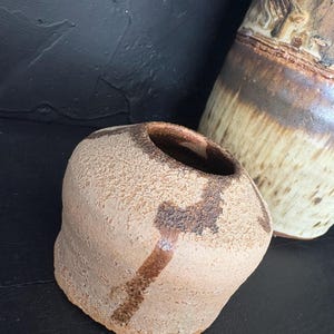 Puede incluir: Un jarrón pequeño, redondo, de cerámica con una superficie texturizada y líneas pintadas de color marrón y negro. El jarrón es de color marrón claro.