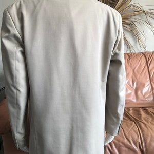 Puede incluir: Un blazer color beige exhibido en un maniqu&iacute;. Se muestra la parte trasera del blazer, destacando su dise&ntilde;o entallado y color neutro. El blazer tiene mangas largas y solapas cl&aacute;sicas.
