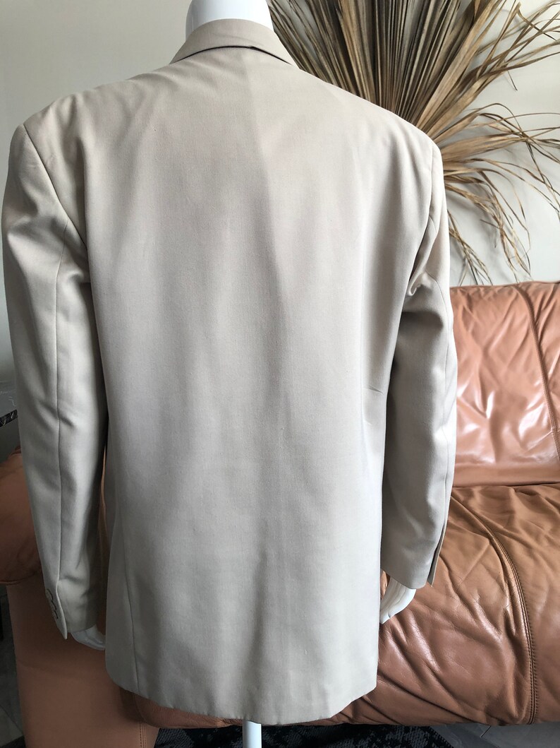 Puede incluir: Un blazer color beige exhibido en un maniqu&iacute;. Se muestra la parte trasera del blazer, destacando su dise&ntilde;o entallado y color neutro. El blazer tiene mangas largas y solapas cl&aacute;sicas.
