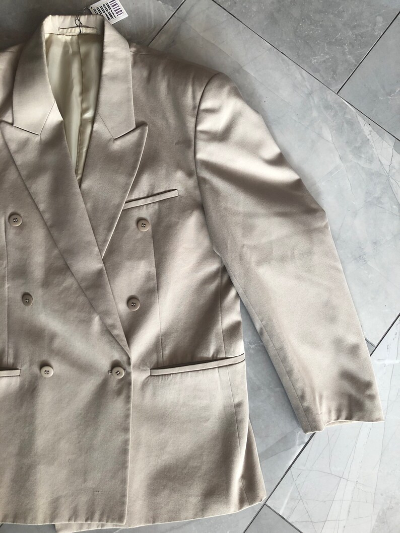 Puede incluir: Un blazer beige de doble botonadura con cuatro botones y dos bolsillos delanteros. El blazer tiene solapas con muescas y un corte entallado. Las mangas son largas y terminan en la mu&ntilde;eca. El blazer est&aacute; hecho de una tela lisa.