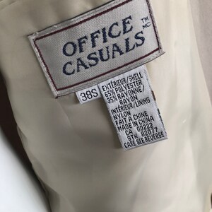 Puede incluir: Primer plano del interior de una chaqueta beige Office Casuals. La etiqueta dice "OFFICE CASUALS" en texto azul marino. Debajo del logotipo hay una etiqueta con la talla "38S" e informaci&oacute;n sobre la composici&oacute;n de la tela en ingl&eacute;s y franc&eacute;s.