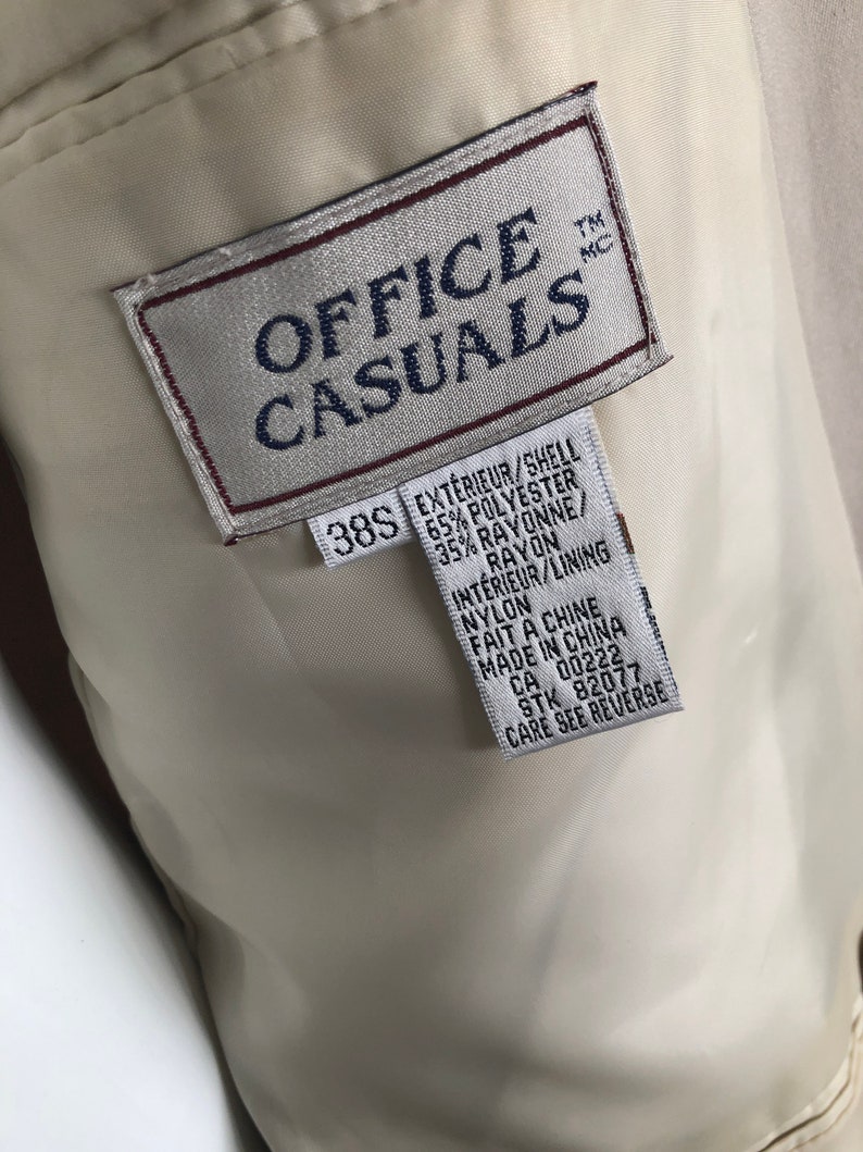 Puede incluir: Primer plano del interior de una chaqueta beige Office Casuals. La etiqueta dice "OFFICE CASUALS" en texto azul marino. Debajo del logotipo hay una etiqueta con la talla "38S" e informaci&oacute;n sobre la composici&oacute;n de la tela en ingl&eacute;s y franc&eacute;s.