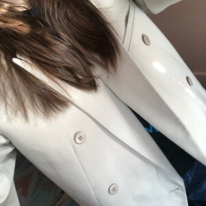 Puede incluir: Una blazer beige claro de doble botonadura con cuatro botones a juego. La blazer tiene un dise&ntilde;o de solapa cl&aacute;sico y se lleva sobre una camiseta negra. Las mangas son largas y la tela parece ser un material liso y tejido.