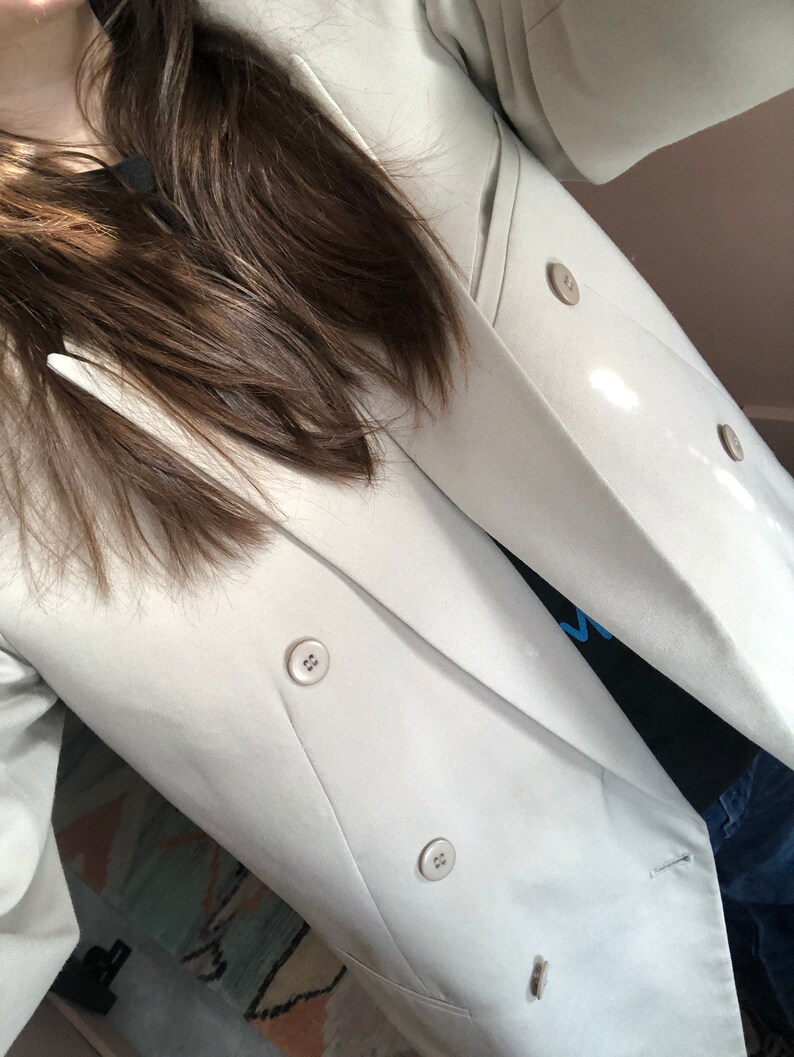 Puede incluir: Una blazer beige claro de doble botonadura con cuatro botones a juego. La blazer tiene un dise&ntilde;o de solapa cl&aacute;sico y se lleva sobre una camiseta negra. Las mangas son largas y la tela parece ser un material liso y tejido.