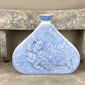 May include: A light blue ceramic vase with a raised design of a person riding a buffalo. The vase has a rounded, triangular shape.