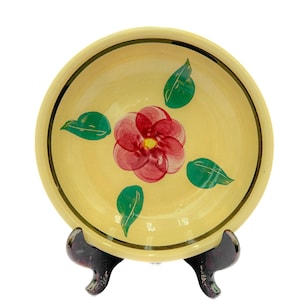 May include: A yellow ceramic bowl with a hand-painted pink flower and green leaves. The bowl has a gold rim and is sitting on a wooden stand.