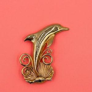 May include: A gold-toned metal brooch shaped like a leaping dolphin. The dolphin is positioned above a textured, fan-shaped seashell base with decorative swirling metal accents on either side, creating a dynamic and fluid aquatic composition.