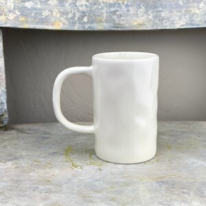 Pu&ograve; includere: Una tazza in ceramica bianca con una superficie leggermente strutturata e un manico arrotondato. La tazza &egrave; appoggiata su una superficie grigio chiaro.