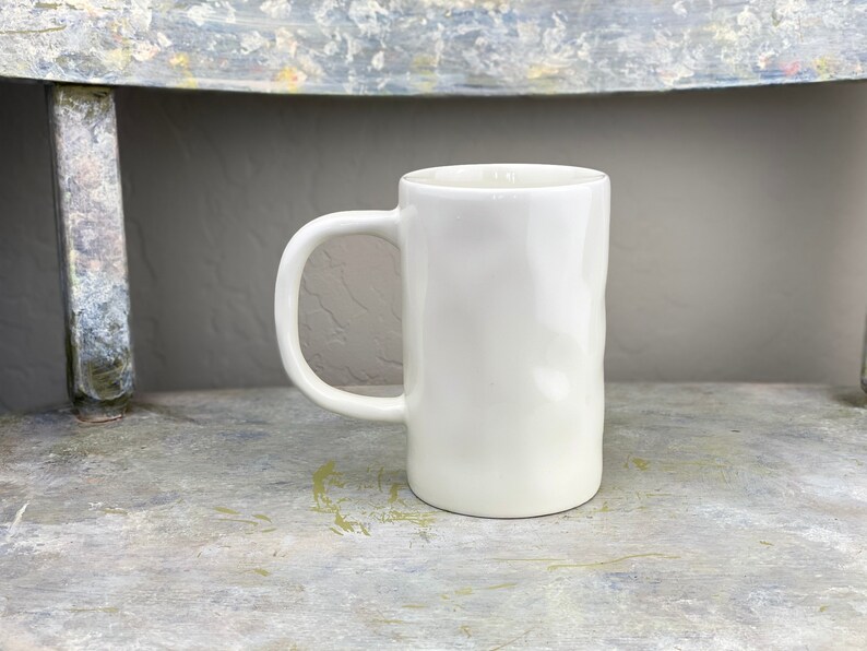 Pu&ograve; includere: Una tazza in ceramica bianca con una superficie leggermente strutturata e un manico arrotondato. La tazza &egrave; appoggiata su una superficie grigio chiaro.